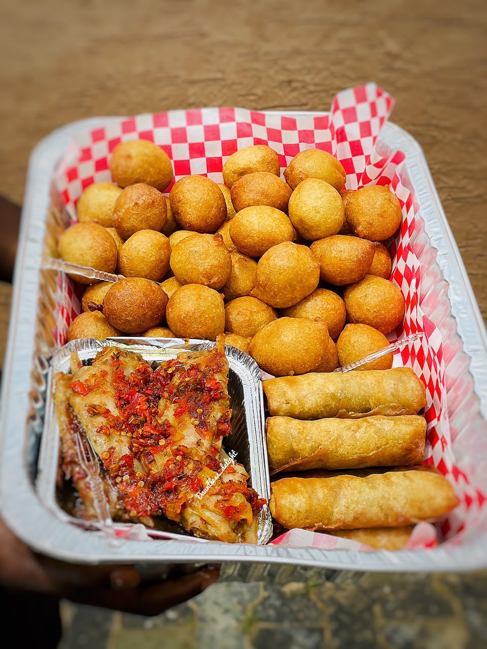 Tray of puff-puff, spring rolls and peppered fish on checkered paper