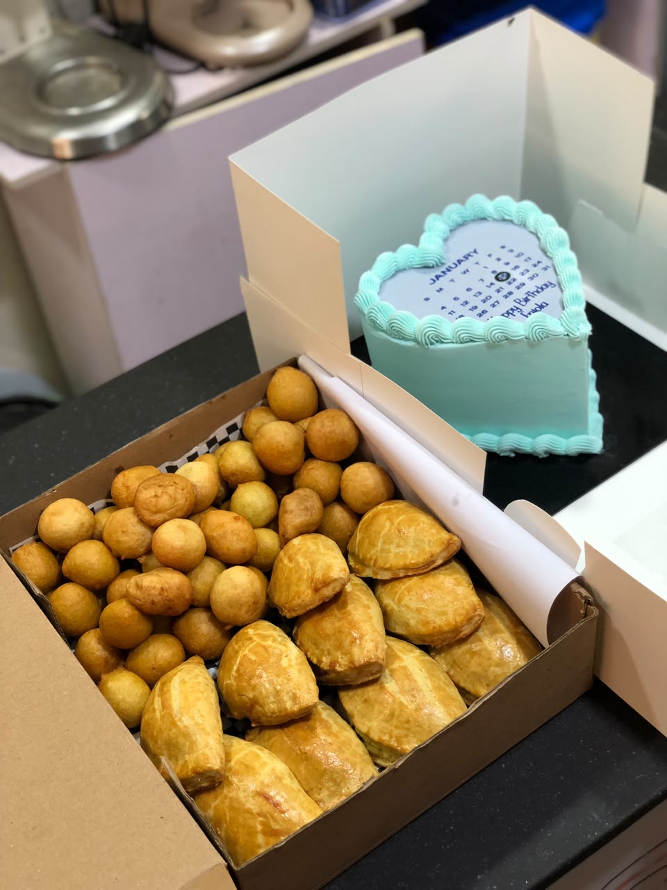 Box of puff-puff and meat pies beside a mint heart shaped birthday cake